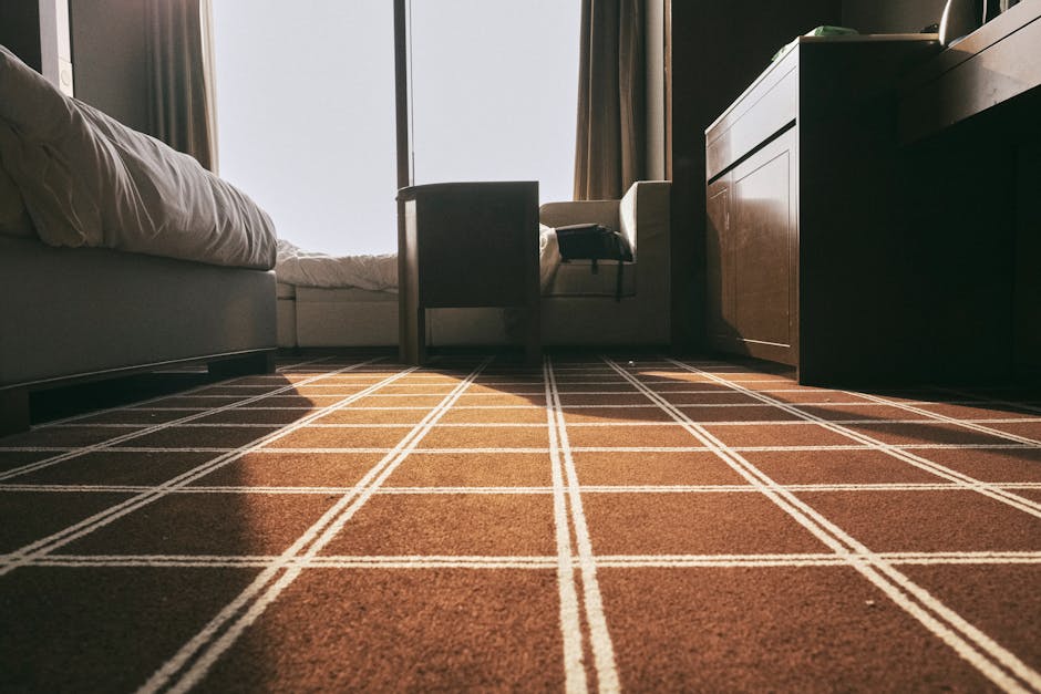 A professional cleaner dressed in full protective white coveralls, including gloves and a mask, is performing deep surface cleaning on a beige, plush carpet in a bright, modern living room. The room f
