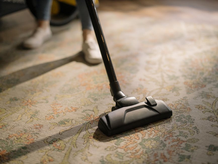 A close-up of a person vacuuming a patterned area rug with a modern, black vacuum cleaner in a well-lit room. The rug features intricate floral designs in soft pastel shades, and the vacuum's head is positioned on the surface, demonstrating surface cleaning. Behind the vacuum, a person wearing white shoes and dark trousers is partially visible, indicating a professional cleaning process. The room appears tidy and bright, emphasizing the thoroughness of the cleaning service provided by Knightsbridge Carpet Cleaners at Eaton Square in Knightsbridge.
