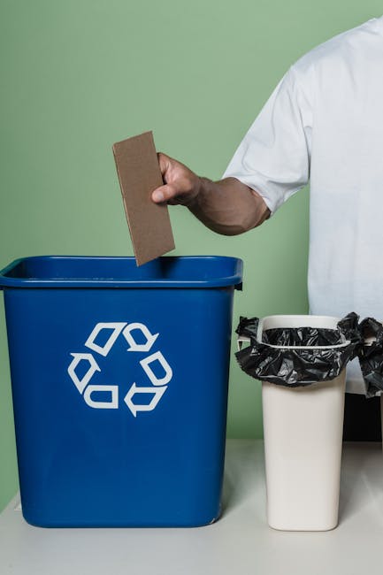 A person wearing a white shirt is holding a brown cardboard scrap over a blue recycling bin with a white recycling symbol, placed on a white surface. To the right, there is a smaller white trash can l