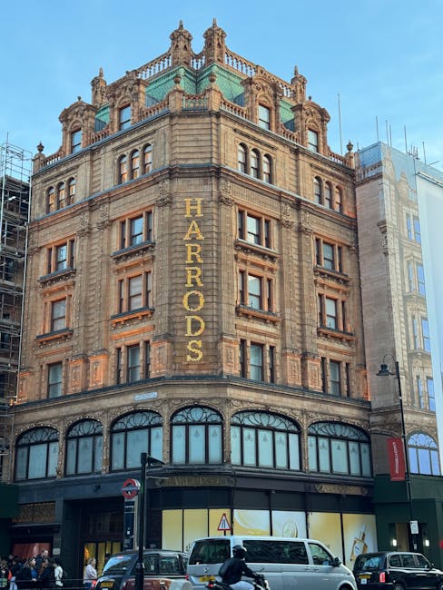 Exterior view of a historic multi-story building in Knightsbridge, London, with ornate architectural details and large arched windows on the ground floor. The building features a prominent vertical sign reading 'HARRADS' on its facade. The lower section appears to house retail spaces with large display windows, while the upper floors are constructed from stone with decorative elements. The street scene includes cars and pedestrians, with a clear blue sky overhead. The image exemplifies urban architecture aligned with professional cleaning standards, highlighting the importance of maintaining building exteriors, which is relevant for services offered by Knightsbridge Carpet Cleaners for deep cleaning and surface maintenance near Harrods on Brompton Road.