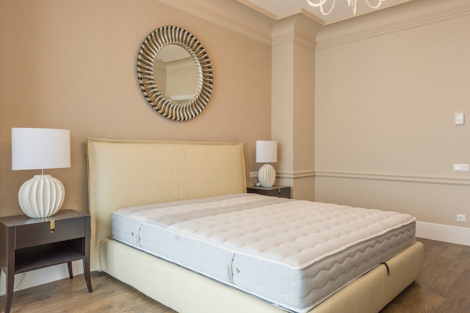 A bedroom featuring a neatly made bed with a cream upholstered headboard and a white mattress, flanked by two dark wooden bedside tables. Each table holds a white ceramic table lamp with a rounded, ri