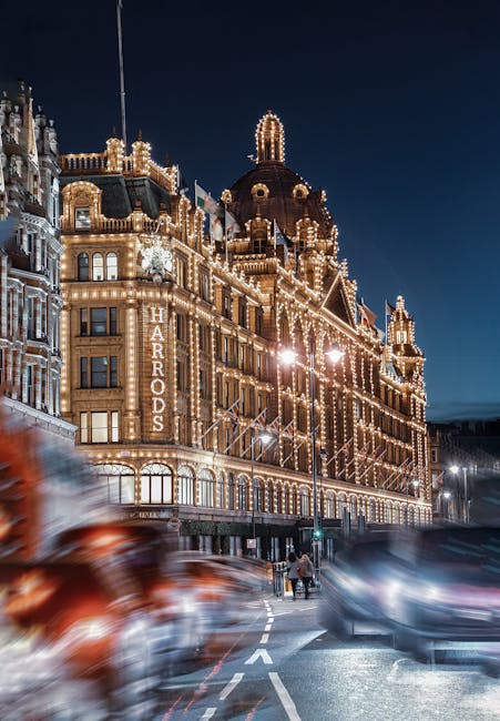 A nighttime view of the historic Harrods department store illuminated with warm, white decorative lighting outlining the building's intricate architectural details, including domes and turrets. The st
